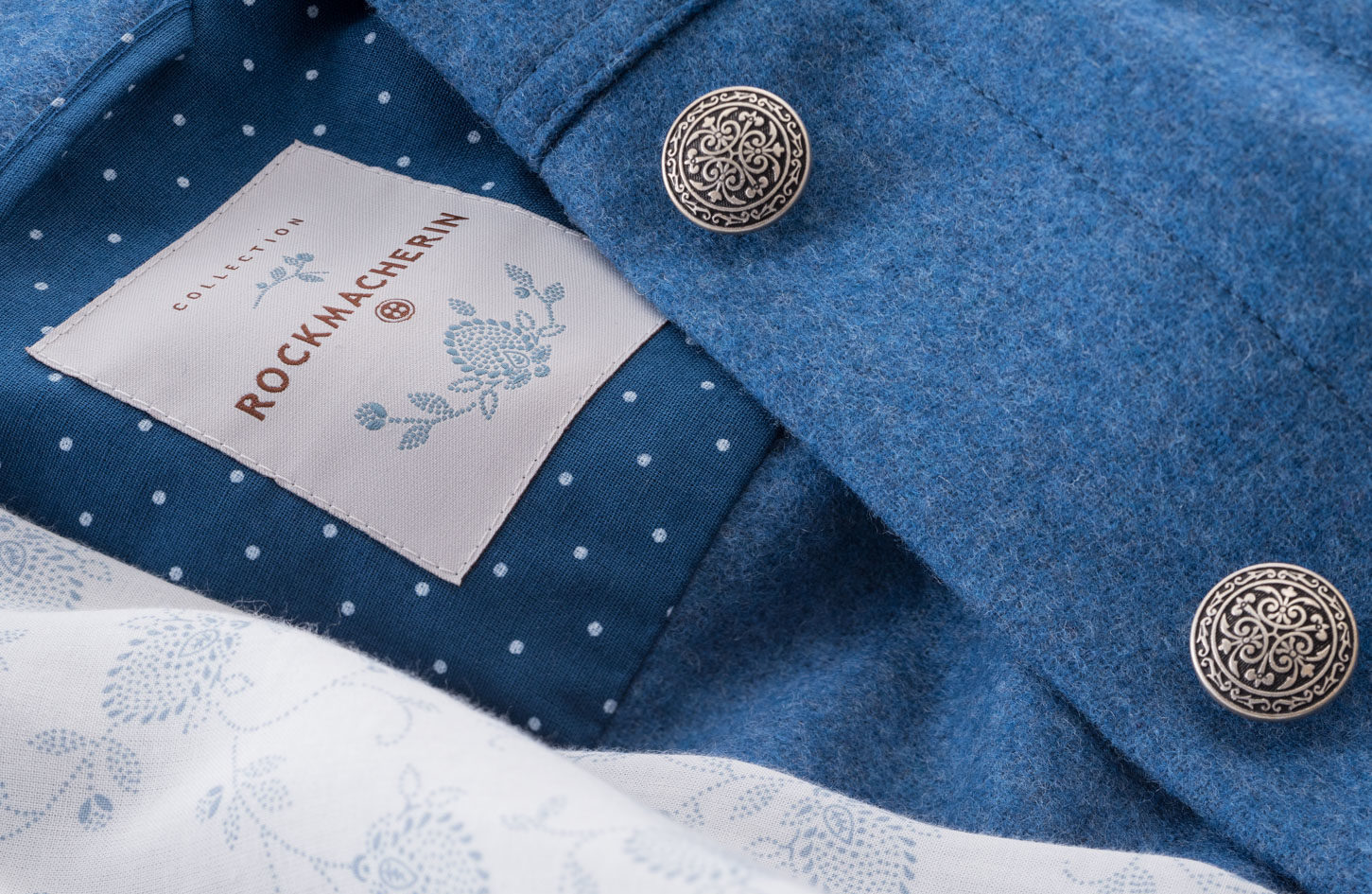 Ein Rockmacherin Kleidungsstück im Anschnitt in einem mittleren Blau, zwei florale Metallknöpfe und das Rockmacherin Etikett