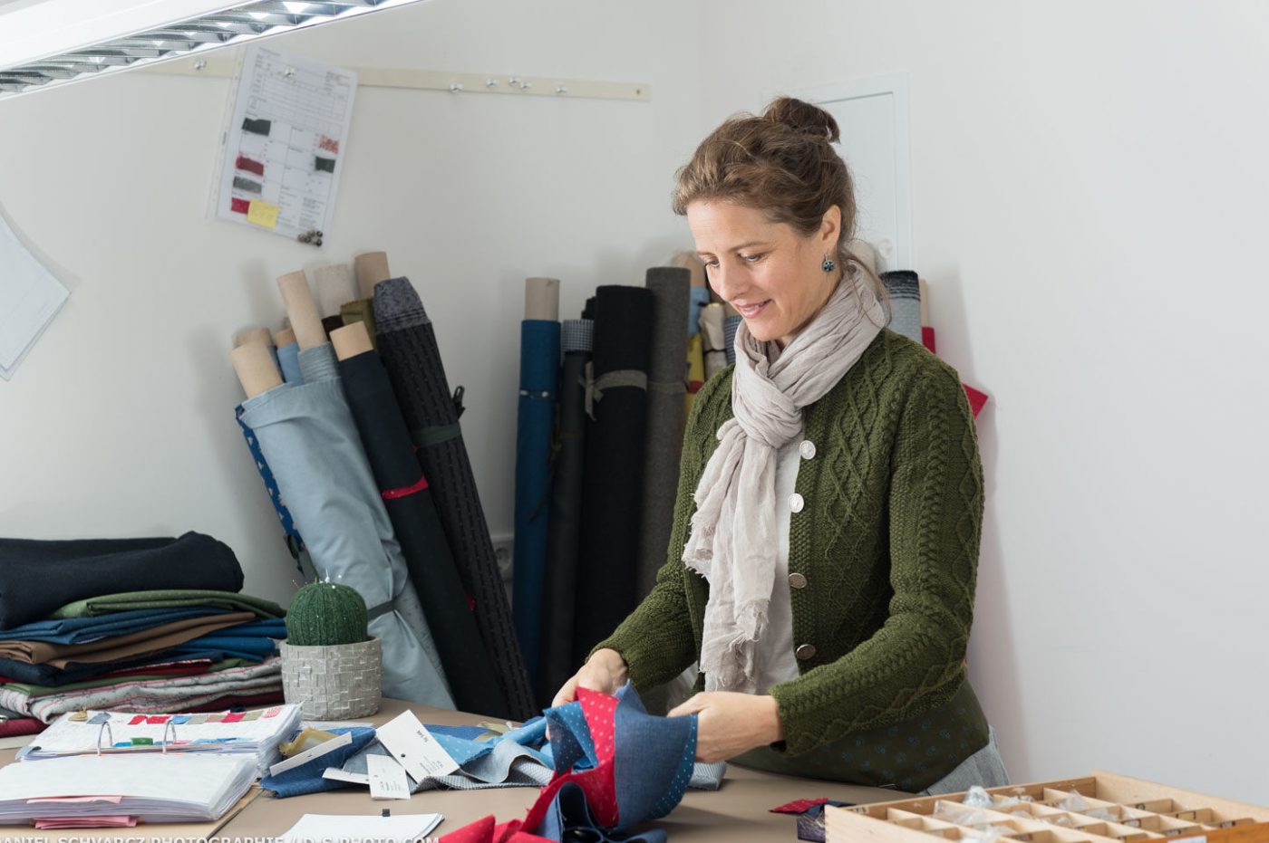 Caroline Lauenstein im Designprozess mit Stoffrollen im Hintergrund am Designtisch mit Stoffelaschen, Designordner und Schneiderzubehör.