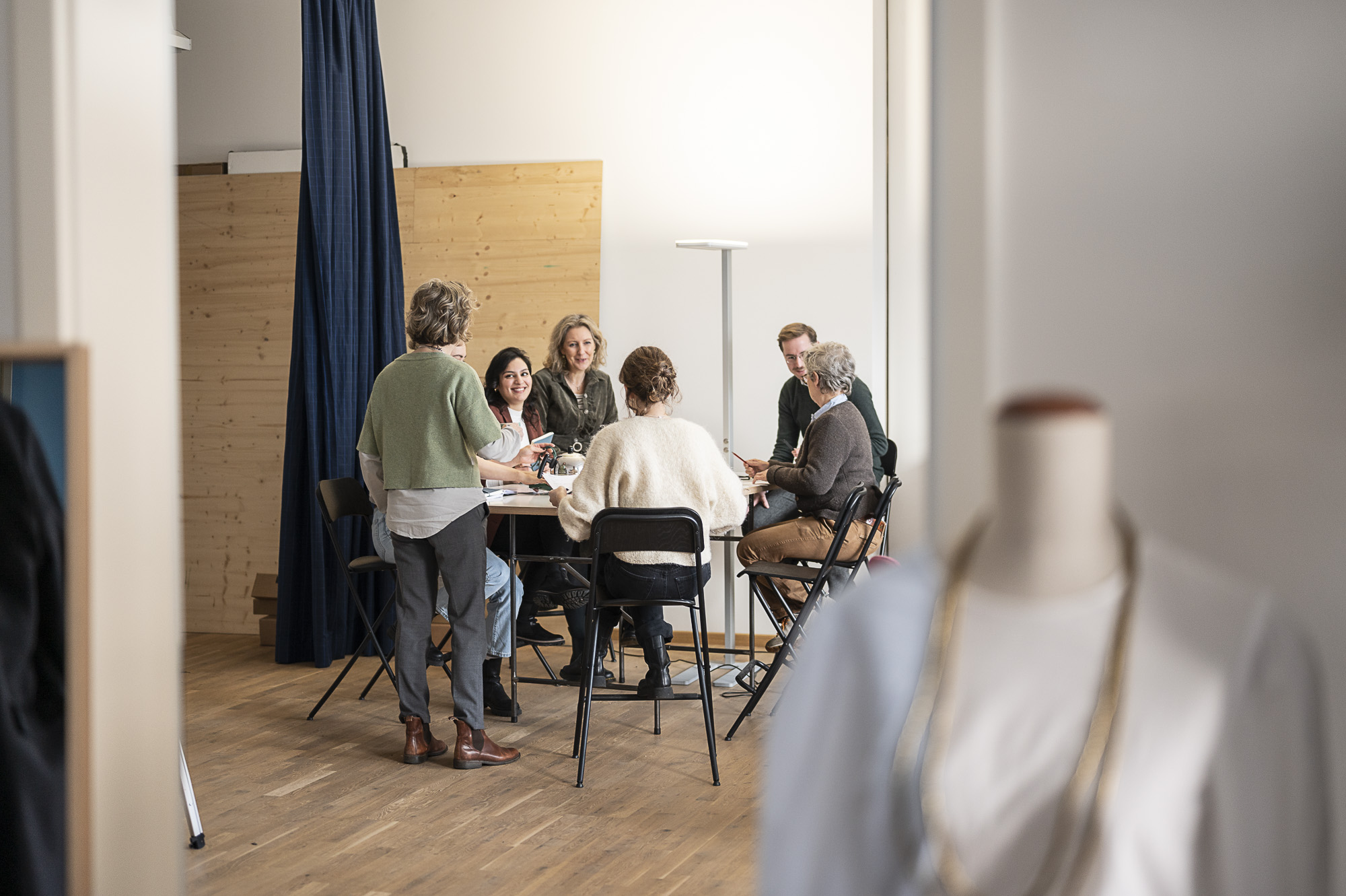 Teamfoto Hinter den Kulissen - zeigt Team und an einem hohen Tisch steht Caroline mit dem Rücken zum Team gewandt das Team sitzend um den Tisch auf hohen Stühlen ausgelassene Stimmung