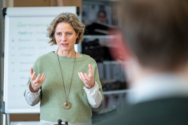 Caroline mit Flipchart im Hintergrund in einem Meeting sprechend und gestikulierend